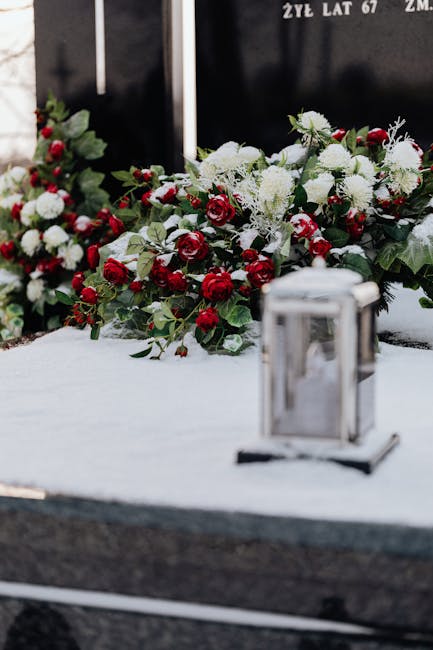 A close-up view of a wooden casket adorned with an array of fresh floral arrangements, including white roses, pale pink peonies, and cream-colored lilies, resting on top and around the sides. The flow