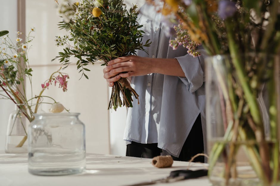 A woman with long, silver braided hair wearing a purple fleece jacket and blue gloves is holding a bouquet of white roses wrapped in clear cellophane, examining the flowers closely at an outdoor marke