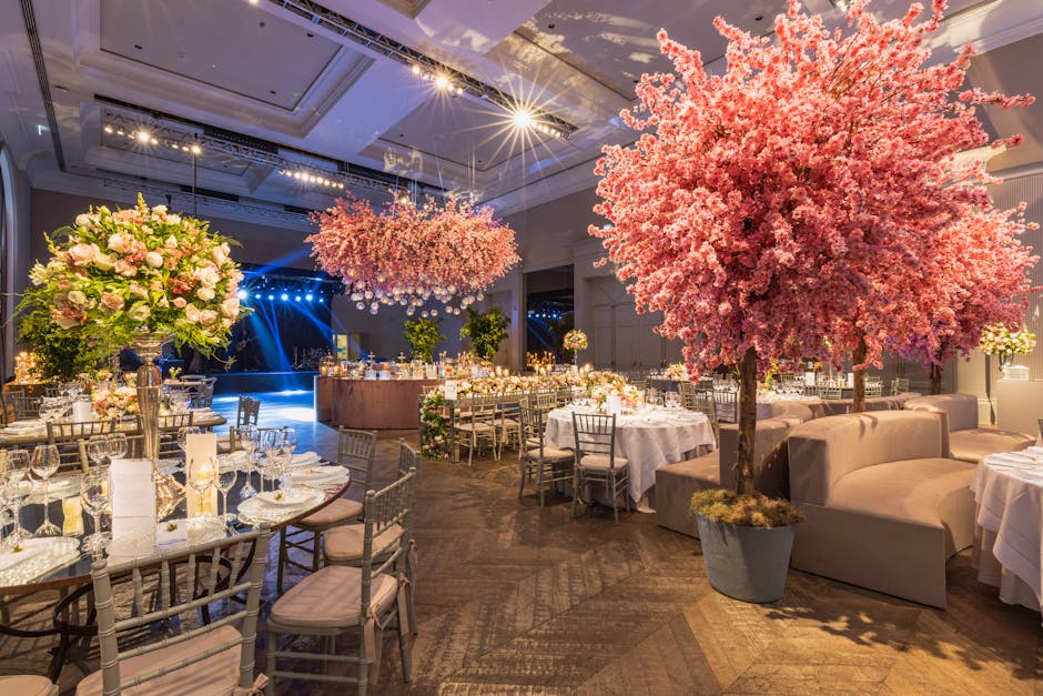 A round banquet table at a formal event set with elegant glassware, white dinner plates, and polished cutlery, arranged beneath a large, lush floral centerpiece consisting of white roses, hydrangeas,