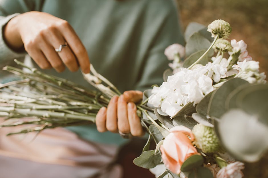A floral arrangement featuring a single large pink and cream rose at the center, surrounded by a dense cluster of small white daisy-like flowers with yellow centers. The bouquet is being held by a per