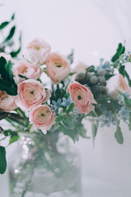 A bouquet of pale peach roses with fully bloomed petals and soft, velvety texture, arranged in a clear glass vase filled with water. The roses are complemented by sprigs of green foliage with small, e