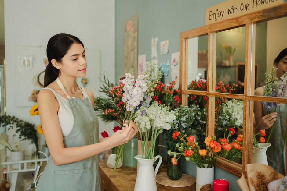 A woman with shoulder-length brown hair and a grey top is arranging a bouquet of fresh flowers in a white ceramic pitcher. The bouquet features pink and white lisianthus with delicate, ruffled petals,